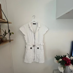 Vit kortärmad manchesterklänning storlek 38 - Snygg vit kortärmad klänning från PrettyLittleThing med dragkedja framtill och ett matchande bälte i midjan. Klänningen har stora fickor med knappar som ger en cool look. Perfekt för en stilren och avslappnad outfit. Manchester material. Fint skick. 