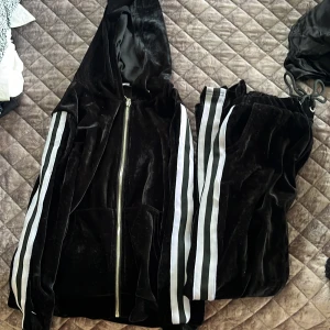 Svart velourjacka och byxor från Born in Stockholm - Snygg svart velourjacka med huva och dragkedja från Born in Stockholm. Jackan har vita ränder längs ärmarna som ger en sportig look. Perfekt för en avslappnad stil.