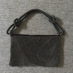 Svart glittrig axelväska - Snygg svart axelväska med glittrande detaljer. Väskan har en unik design med ett flätat axelband och en praktisk dragkedja för säker förvaring. Perfekt för att ge en extra touch till din outfit.