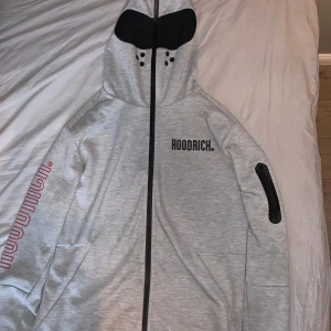 Unik zip hoodie från Hoodrich - Säljer en grå huvtröja från Hoodrich med dragkedja och tryck i svart och rött. Tröjan har en luva och långa ärmar med tryck längs ena ärmen. Perfekt för en casual look.