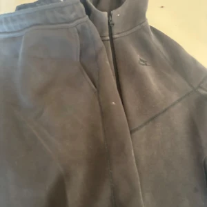 Grå fleecejacka från Nike - Säljer en grå fleecejacka från Nike med dragkedja och huva. Jackan har långa ärmar och två fickor med dragkedja framtill. Perfekt för kyliga dagar.