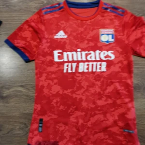 Röd fotbollströja från Adidas - Säljer en röd fotbollströja från Adidas med Olympique Lyonnais-logga och 'Emirates Fly Better' tryck på framsidan. Tröjan har korta ärmar med blå detaljer och ett subtilt mönster över hela tyget. Perfekt för fotbollsfans!