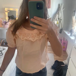 Orange  blus med spetsdetaljer - Säljer en söt orange blus med korta ärmar och vacker spetsdetalj runt halsringningen. Perfekt för en somrig look. Blusen har en lös passform och knytband vid halsen.