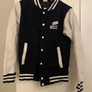 Blå och vit jacka från NOCCO - Snygg blå och vit jacka från NOCCO med vita ärmar och randiga detaljer. Jackan har knappar framtill och två fickor. Perfekt för en sportig look!