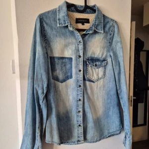 Jeansskjorta från River Island - Snygg jeansskjorta från River Island i en ljusblå tvätt. Skjortan har långa ärmar och två bröstfickor med knappar. Perfekt för en avslappnad stil.