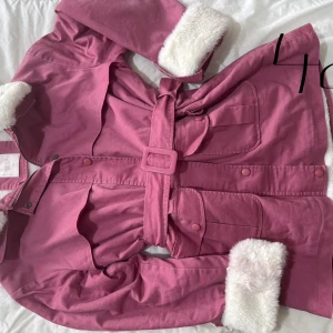 Rosa jacka med vit fuskpäls - Säljer en snygg rosa jacka från Manto Bird med vit fuskpäls på krage och ärmslut. Jackan har ett bälte i midjan och stängs med knappar. Perfekt för kyliga dagar!