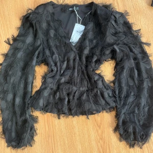 Svart topp med fjäderdetaljer - Snygg svart blus från ginatricot i storlek 36 med prislapp kvar säljer då den aldrig kommit till användning!