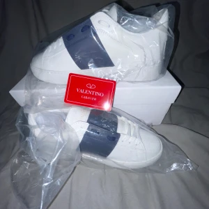 Valentino Garavani sneakers i vitt och blått - Snygga Valentino Garavani sneakers i vitt med en bred blå rem över ovansidan. Skorna har en rund tå och är tillverkade i skinn. Perfekta för en stilren look. Priset kan diskuteras vid snabb affär 💯