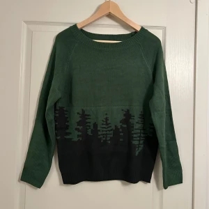 Grön stickad tröja med skogsmotiv - Säljer en grön stickad tröja med ett coolt skogsmotiv i mörkare nyanser. Tröjan har långa ärmar och en rund halsringning. 