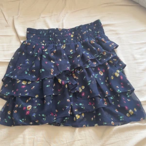 Blommig kjol  - Söt blommig kjol från American Eagle Outfitters i mörkblått med volanger. Perfekt för en somrig look med sitt färgglada blommönster. Elastisk midja för bekväm passform.