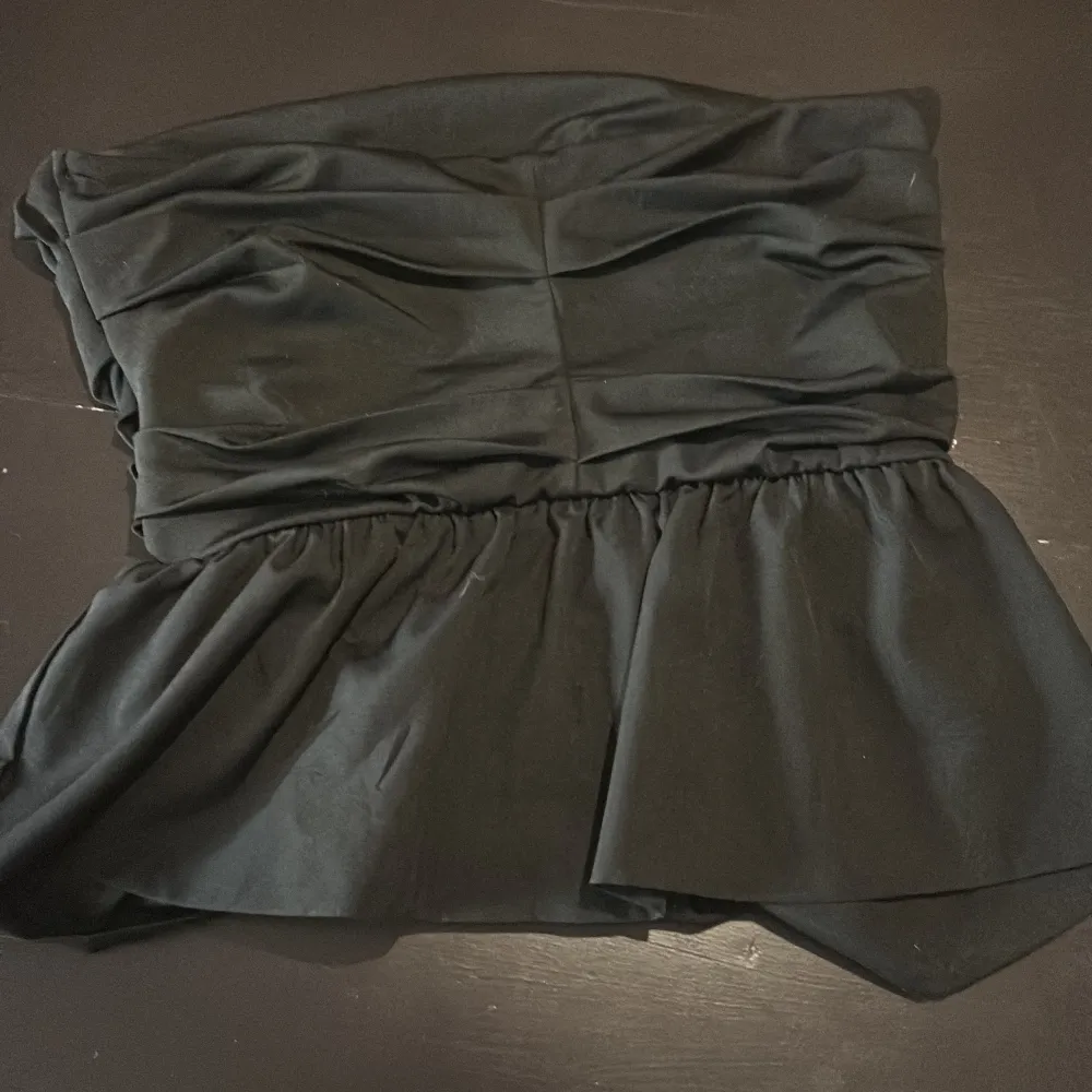 Snygg svart axelbandslös topp med draperad design och volangdetaljer. Perfekt för en stilren och elegant look. Passar bra till både jeans och kjol.. Yläosat.