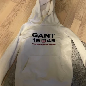 Vit hoodie från GANT - Säljer en vit hoodie från GANT med tryck framtill som visar 'GANT 1949 American Sportswear'. Tröjan har en klassisk känguruficka och en bekväm huva. Perfekt för en avslappnad stil. Fint skick✔️Storlek 146-152