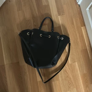 Svart bucket bag från H&M - Stilren svart bucket bag från H&M med guldfärgade metalldetaljer och snörning upptill. Väskan har både handtag och lång axelrem, perfekt för att bära på olika sätt. Tillverkad i syntetmaterial med lätt struktur och rymligt innerfack.