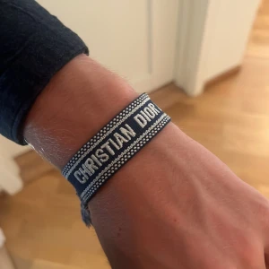 Christian Dior blått tygarmband - Snyggt blått armband från Christian Dior med vit brodyr och texten 'CHRISTIAN DIOR' samt 'J'ADIOR'. Armbandet är gjort i tyg och har justerbara tofsar i ändarna för en cool och avslappnad look.