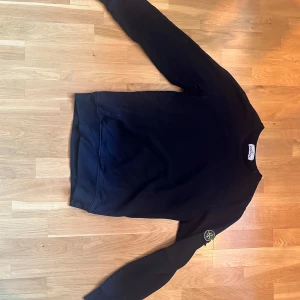 Svart sweatshirt från Stone Island - Svart sweatshirt från Stone Island med klassisk rund hals och lång ärm. Ikonisk logopatch på vänster ärm.