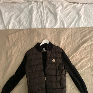 Svart Moncler pufferjacka med stickade ärmar - Säljer en svart pufferjacka från Moncler med quiltad front och stickade ärmar. Jackan har dragkedja framtill och klassisk Moncler-logga på bröstet. Perfekt för dig som vill ha en stilren och sportig look med premiumkänsla. Den har en liten skada där med den halvt avrivna moncler lappen inuti jackan som inte syns när man har på sig jackan därför säljer jag för inte så dyrt, hör gärna av er mvh Douglas