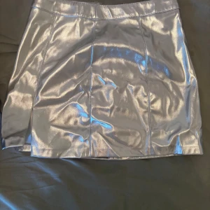 Silvrig kortkjol från Pinkie - Supersnygg silvrig kortkjol från Pinkie med glansig finish och en liten slits bak. Kjolen har en figurnära passform och är perfekt för dig som vill sticka ut. Tillverkad i syntetmaterial som ger en cool metallic look.