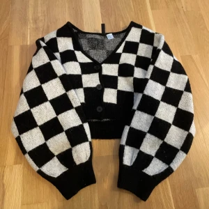 Svartvit rutig kofta med knappar - Snygg croppad kofta med svartvitt rutigt mönster och v-ringning. Koftan har tre knappar framtill, långa ärmar och ribbade muddar. Perfekt för dig som gillar statement-plagg och vill sticka ut lite extra.