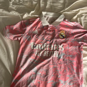 Rosa Real Madrid x Adidas fotbollströja - Snygg rosa Real Madrid fotbollströja från Adidas med grått drakmönster över hela tröjan. Klassisk vit krage, korta ärmar och broderad klubbmärke på bröstet. Materialet är lätt och andas, perfekt för träning eller match.