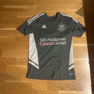 Houston Dynamo grå fotbollströja Adidas - Snygg grå fotbollströja från Houston Dynamo med vita detaljer och Adidas-logga på bröstet. Tröjan har korta ärmar, tryckt sponsor på framsidan och stort nummer på ryggen. Tillverkad i lätt och ventilerande material, perfekt för träning eller match. Diskuterbart pris 