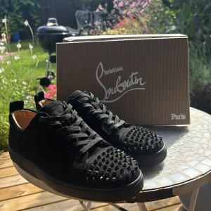 Christian Louboutin svarta sneakers med nitar - Svarta sneakers från Christian Louboutin i mocka med ikoniska röda sulor. Skorna har snörning och är dekorerade med svarta nitar över tån för en edgy look. Insidan är i ljus skinn och sulan är platt. Perfekta för dig som vill sticka ut. Skorna är använda 1 gång!
