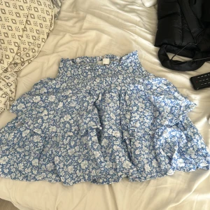 Blåvit blommig volangkjol - Supersöt kort kjol med blått och vitt blommönster och volanger. Resår i midjan gör den bekväm och lätt att bära. Perfekt för varma dagar när du vill känna dig extra fin.