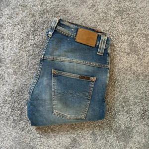 Blå jeans från Nudie Jeans, 29/34 - Snygga blå jeans från Nudie Jeans med klassisk femficksdesign och orangea sömmar. Jeansen har en lätt tvättad look och är tillverkade i bomull. Passar dig som gillar en tidlös och avslappnad stil.