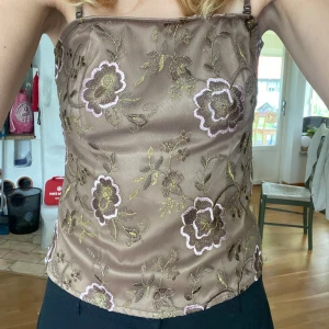 Brun blommig linnetopp med broderi - Supersöt brun linnetopp från Vila i storlek M. Tunna axelband och broderade blommor i rosa och grönt. Toppen har en lätt glansig yta och är figurnära. 