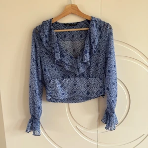 Blå mönstrad blus med volang - Supersnygg blå blus från Zara med grafiskt mönster och volang vid v-ringningen. Långa, transparenta ärmar med resår vid handlederna och croppad passform. 