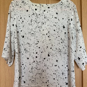 Vit blus med svart prickigt mönster - Vit blus från Stockh LM i storlek 40 med svart prickigt mönster som ser ut som färgstänk. Blusen har trekvartsärmar och en liten knapp i nacken. Luftigt material och rak passform, perfekt för en cool och stilren look.