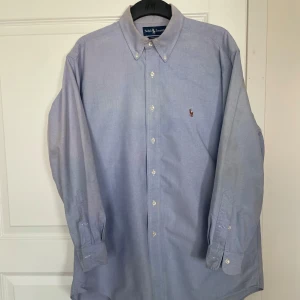 Ljusblå skjorta från Ralph Lauren xl - Klassisk ljusblå skjorta från Ralph Lauren med button-down krage och broderad logga på bröstet. Tillverkad i 100% bomull oxford, långärmad och med vita knappar framtill. Perfekt för en clean och stilren look.