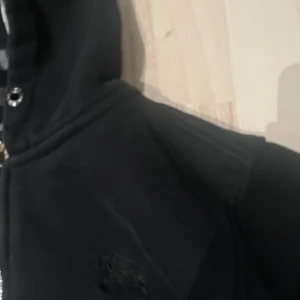 Svart Burberry hoodie med dragkedja - Svart hoodie från Burberry med klassiskt rutigt foder i huvan. Tröjan har dragkedja framtill, två fickor och broderad logga på bröstet. Perfekt för dig som gillar stilrena och exklusiva streetwear-plagg.