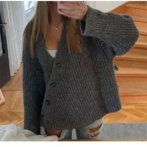 Grå asymmetrisk stickad kofta från & Other Stories - Mysig grå kofta från & Other Stories med oversized passform och bred ribbstickning. Koftan har v-ringning, knappar framtill och långa ärmar. Perfekt att slänga över en t-shirt eller linne för en chill look.