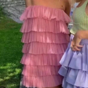 Rosa volangklänning axelbandslös - Superfin rosa kortklänning med flera lager volanger och plisserat tyg. Axelbandslös modell som ger en cool och lekfull vibe, perfekt för dig som vill sticka ut. Klänningen har en loose passform och är lätt och luftig.