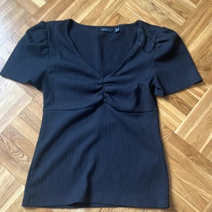 Svart v-ringad topp från Vero Moda - Svart topp från Vero Moda med v-ringning och korta puffärmar. Toppen har snygg rynkdetalj framtill och är tillverkad i ett ribbat material som ger en cool struktur. Perfekt för dig som gillar stilrena plagg med en twist.