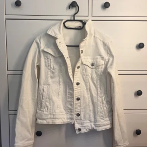 Vit jeansjacka med silvriga knappar - Snygg vit jeansjacka med klassisk krage och silvriga knappar framtill. Jackan har långa ärmar, bröstficka med knapp och två sidofickor. Perfekt att slänga över en hoodie eller t-shirt för en clean och fräsch look.