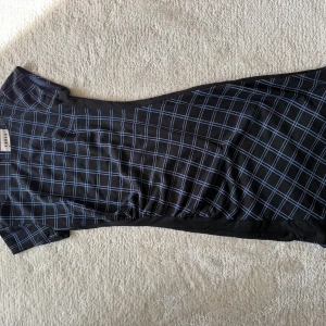 Svart rutig klänning Karen by Simonsen - Snygg svart långklänning från Karen by Simonsen med blått rutmönster. Klänningen har korta ärmar, figurnära passform och dragkedja i ryggen. Perfekt för dig som gillar stilrena och moderna plagg med en twist.