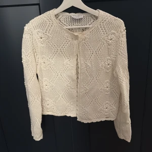 Vit stickad kofta från Holly & Whyte - Supersöt vit stickad kofta från Holly & Whyte med rund halsringning och ett diskret mönster i strukturen. Långärmad och med en knapp framtill. Perfekt att slänga över en topp för en chill och stilren look.