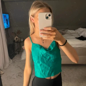 Grön satin linnetopp med tunna band - Snygg grön linnetopp i satin med tunna axelband. Croppad modell, perfekt för varma dagar. Materialet är glansigt. Aldrig använd 