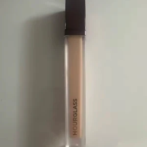 Hourglass flytande concealer - Flytande concealer från Hourglass i en stilren, genomskinlig tub med svart lock. Färgen är ljusbeige och formulan är krämig, perfekt för att täcka mörka ringar eller blemmor. Snygg minimalistisk design med Hourglass-loggan tryckt på tuben.