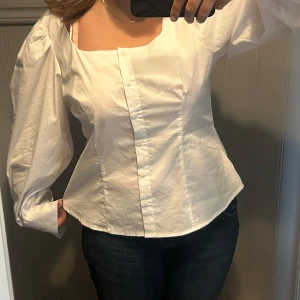 Vit skjorta med puffärm och knappar - Säljer en vit skjorta med fyrkantig urringning och puffiga långärmar. Skjortan har figurnära passform och knäppning framtill med dolda knappar. Perfekt för dig som gillar en stilren men trendig look.