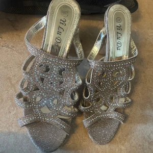 Glittriga silvriga sandaler med klack - Snygga sandaler i silver med glittriga stenar och öppen tå. Skorna har en dekorativ utskuren design och bred klack som ger stabilitet. Perfekta för dig som vill sticka ut med extra bling och glam. Materialet är syntet och de har en elegant, festlig känsla.