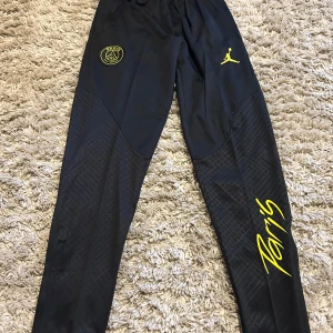 Svarta PSG x Jordan träningsbyxor - Snygga svarta träningsbyxor från PSG x Jordan med gula detaljer och loggor. Byxorna har smal passform, elastisk midja och coolt mönstrade partier på benen. Perfekta för fotboll eller chill. Paris-tryck i gult längs benet ger extra stil.