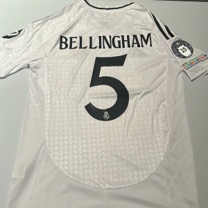 Real Madrid Bellingham #5 matchtröja - Snygg vit Real Madrid fotbollströja från Adidas med Bellingham och nummer 5 på ryggen. Tröjan har svarta detaljer, klubbmärke, Champions League-märke och FIFA-märke på bröstet. Tillverkad i lätt funktionsmaterial med korta ärmar och subtilt mönster i tyget. Replika. Endast handtvätt gäller!