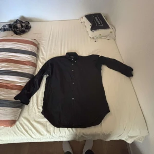 Svart skjorta Polo Ralph Lauren L - Klassisk svart skjorta från Polo Ralph Lauren i storlek L. Skjortan är i 100% bomull, har långa ärmar, knäppning framtill och ikonisk röd broderad logga på bröstet. Perfekt för dig som gillar stilrena och tidlösa plagg.