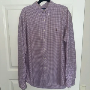 Lila rutig skjorta från Ralph Lauren - Snygg lila och vit rutig skjorta från Ralph Lauren i klassisk passform. Skjortan har button-down krage, långa ärmar och broderad logga på bröstet. Tillverkad i mjuk bomull och stängs med vita knappar framtill. Passar en som är emellan 175-185. OBS, fläck längst ned på baksidan.