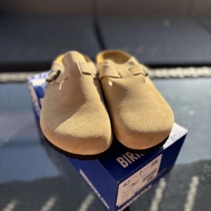 Birkenstock Boston Khaki mocka 42 - Birkenstock Boston i khaki mocka med klassisk slip-in design och justerbart spänne i metall. Skorna har en mjuk ovandel i mocka och en bekväm korksula. Perfekta för chill dagar och enkel stil.