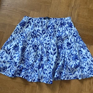 Blåvit mönstrad kjol från Gina Tricot stl M - Supersöt kort kjol från Gina Tricot med blått och vitt mönster. Kjolen har en luftig och volangig design med resår i midjan, perfekt för varma dagar. Tillverkad i mjuk bomull som känns skön mot huden. Snygg att styla med en enkel topp och sneakers.