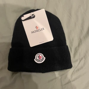Svart Moncler mössa i ull - Svart ribbstickad mössa från Moncler med klassisk uppvikt kant och broderad Moncler-logga framtill. Tillverkad i mjuk ull som håller dig varm och snygg under kyliga dagar. Perfekt accessoar för en stilren look.