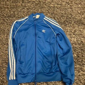 Blå Adidas tröja med vita ränder - Snygg blå vindjacka från Adidas med klassiska vita ränder längs ärmarna och dragkedja framtill. Jackan har hög krage, två fickor och Adidas-logga på bröstet. Perfekt för en sportig och avslappnad stil.